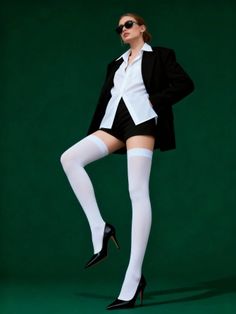Woman in a black blazer, white shirt, shorts, and thigh highs posing against a green background