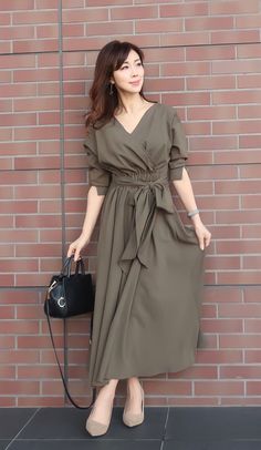 Woman in a long olive green dress with a black handbag standing against a brick wall