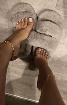 Feet with white nail polish and anklets, wearing fluffy gray slippers indoors