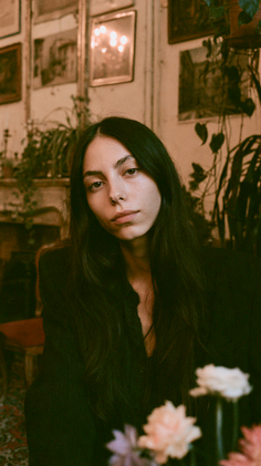 Woman with long dark hair in an indoor setting with plants and warm lighting