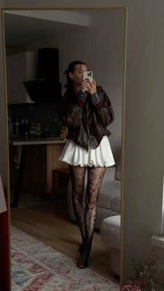Woman in a brown leather jacket and white skirt taking a mirror selfie