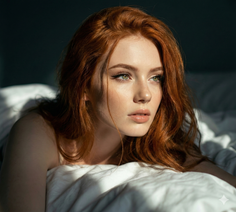 Woman with red hair and green eyes lying in bed with soft lighting