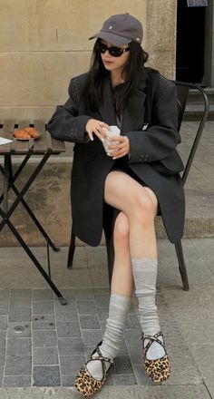 Woman in a blazer, baseball cap, and sunglasses sitting outdoors with exposed legs
