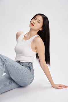 Woman in a white tank top and blue jeans sitting on a white background