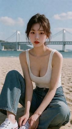 Woman in a white tank top and jeans sitting on a sandy beach with a bridge in the background