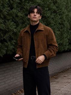 Man in a brown jacket and black turtleneck holding a phone outdoors