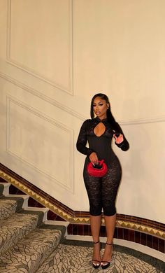 Woman in a black lace jumpsuit and high heels holding a red handbag