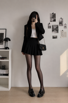 Woman in a black blazer and pleated skirt taking a mirror selfie indoors