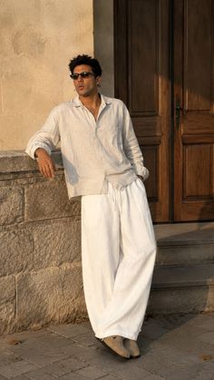 Man in a linen shirt and white pants leaning against a stone wall at golden hour