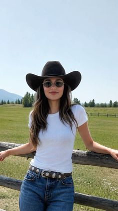 Woman in a cowboy hat and sunglasses leaning on a wooden fence in a green field