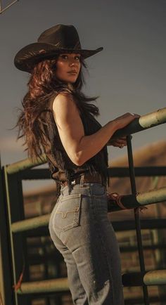 Woman in a cowboy hat and fringe vest leaning on a fence outdoors