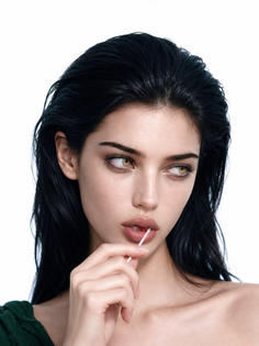 Woman with long wet black hair and green eyes holding a pink stick to her lips