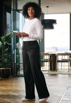 Woman with curly hair in a white shirt and black pants holding a tablet indoors