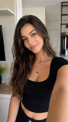 Woman with long dark hair wearing a black top and necklace, taking a selfie indoors