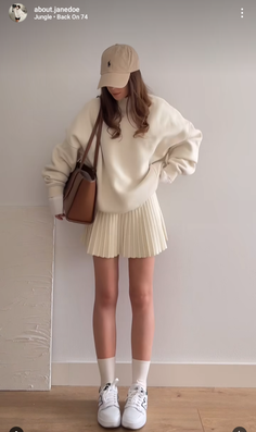 Woman in a cream sweatshirt and pleated skirt with a baseball cap and sneakers