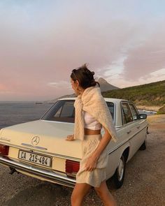 Woman in a knitted set leaning on a vintage car at sunset by the ocean