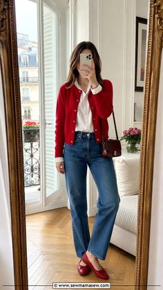Woman in a red cardigan and blue jeans taking a mirror selfie indoors