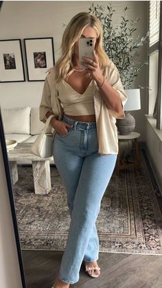 Woman in light wash jeans and a crop top taking a mirror selfie indoors