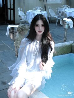 Woman with long black hair in a white dress sitting by blue water at an outdoor cafe