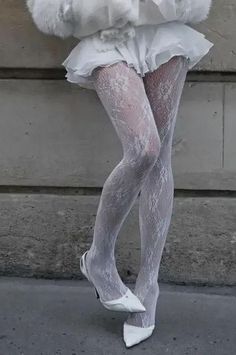 Person wearing white lace tights, a white skirt, and white heels against a concrete wall