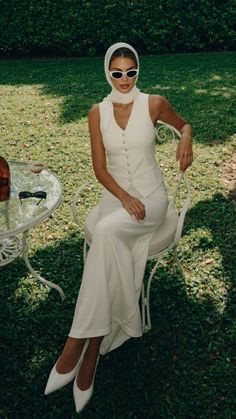 Woman in a white outfit with a head scarf and sunglasses sitting in a garden