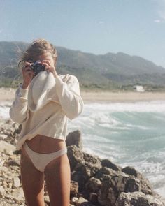 Woman in a bikini and sweater taking a photo on a rocky beach
