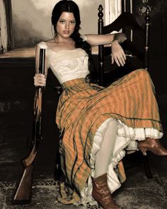 Woman with a shotgun, wearing a long skirt and corset top, sitting indoors