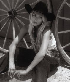 Woman in a cowboy hat and tank top sitting outdoors next to wagon wheels
