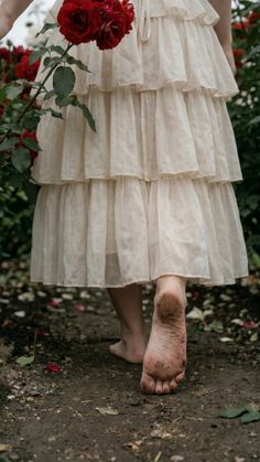 Person in a ruffled dress walking barefoot through a garden with red roses