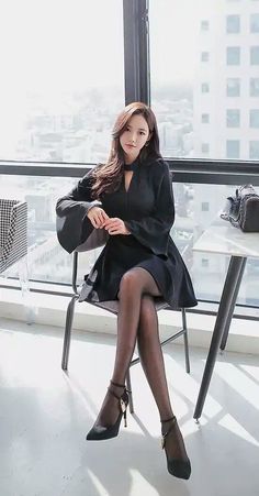 Woman in a black dress and pantyhose sitting by a window overlooking a cityscape