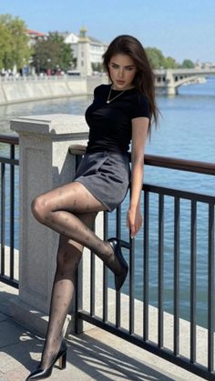 Woman in a black top, short skirt, and sheer tights posing by a river