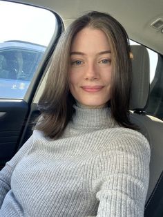 Woman with brown hair and blue eyes wearing a grey turtleneck in a car