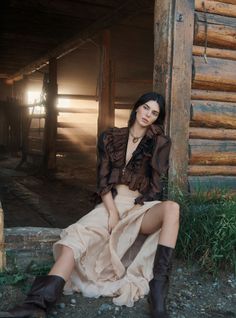 Woman in a ruffled top and long skirt sitting by a wooden cabin