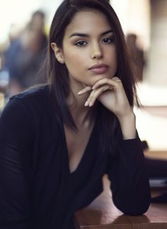 Woman with dark hair in a black deep v-neck shirt, resting her chin on her hand