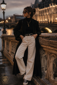Woman in a long black coat and white pants leaning on a bridge railing at night