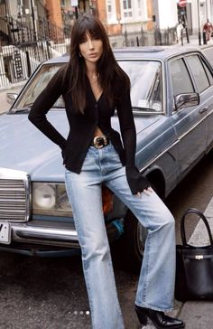 Woman in a black cardigan and light wash jeans leaning on a car on a street