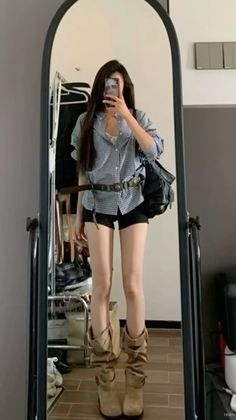 Woman in a gingham shirt, black shorts, and cowboy boots taking a mirror selfie