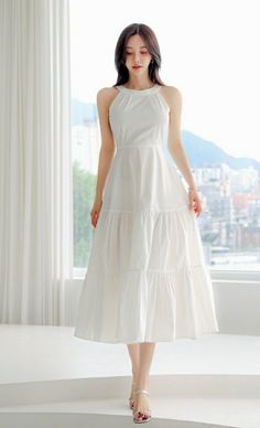 Woman in a white halter neck dress standing by a window with a cityscape view
