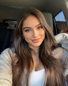 Woman with long brown hair wearing a white tank top and cardigan in a car