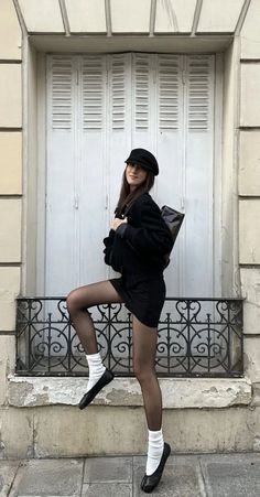 Woman in a black dress, tights, and hat posing outdoors against a building