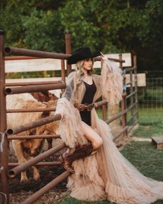 Woman in a cowboy hat and ruffled dress posing by a farm fence with cattle