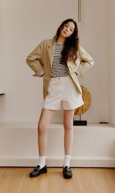 Woman in a khaki blazer, striped shirt, and white shorts posing indoors