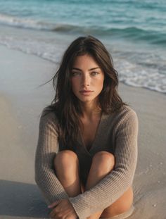 Woman in a sweater sitting on a sandy beach with waves in the background