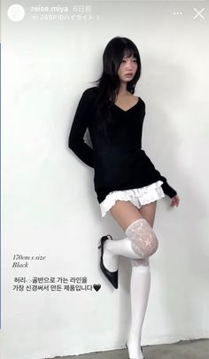 Woman in a black top, white shorts, and lace stockings posing against a white wall