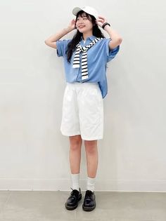 Woman in a blue shirt, white shorts, and a baseball cap posing against a white wall