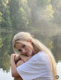 Blonde woman in a white t-shirt posing by a lake with a forest in the background