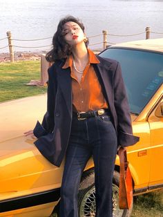 Woman in a dark blazer and orange shirt leaning on a yellow car at golden hour