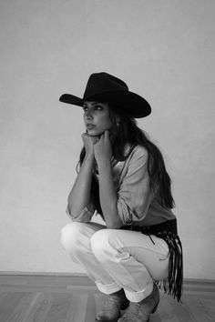 Woman in a cowboy hat and denim shirt squatting against a plain wall