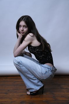 Young woman with long dark hair in a black lace top and jeans, squatting on a wooden floor