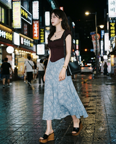 Woman in a dark top and patterned midi skirt walking on a wet city street at night